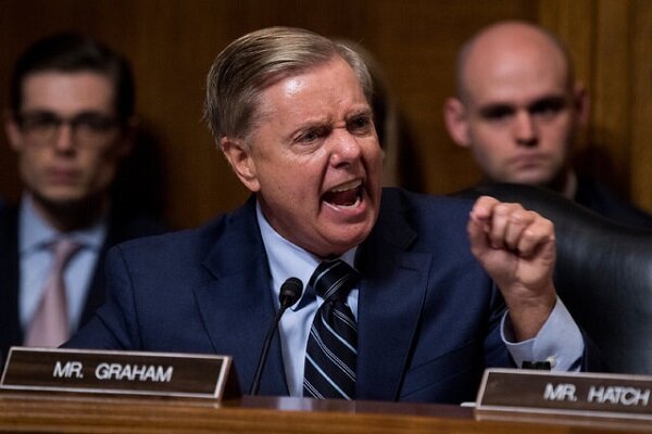 A man in a suit gestures emphatically while speaking at a wooden dais labeled "MR. GRAHAM," with two other men seated behind him.