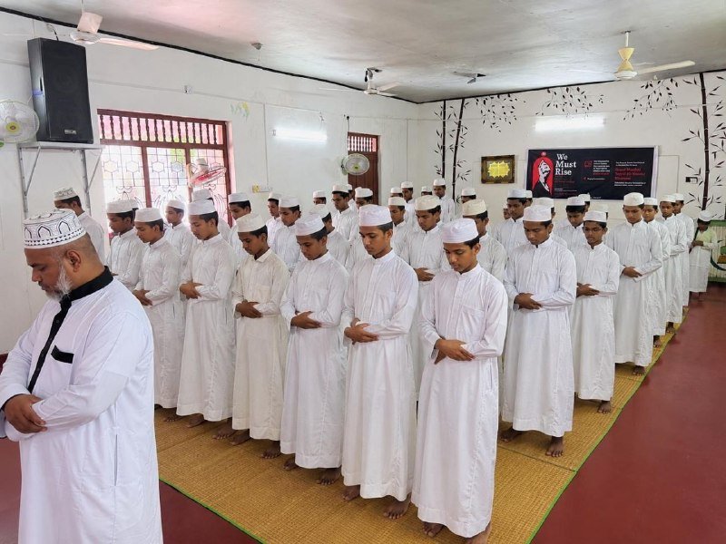 Rows of men and boys in white robes and caps stand barefoot on matting with hands folded, facing the same direction in a room with a "We Must Rise" poster.