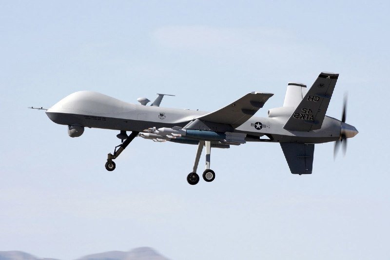 A gray military drone with "HS" and "U.S. Customs" markings flies in a clear sky, its landing gear extended beneath mountainous terrain.