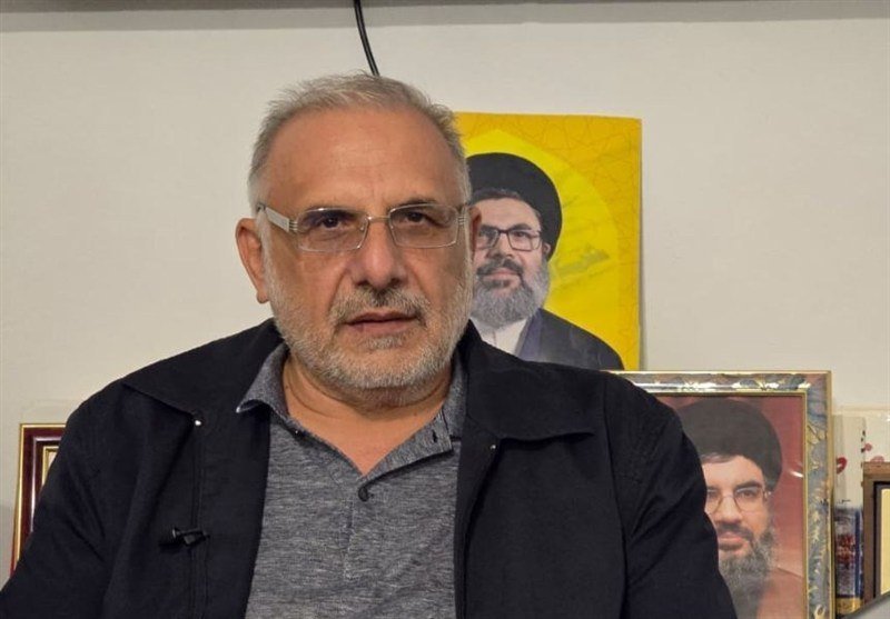 A bearded man with glasses speaks on camera indoors, with framed portraits of clerical figures hanging on the wall behind him.