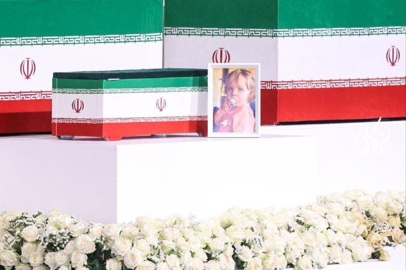 A flag-draped casket and a framed portrait of a young child are displayed on a white platform surrounded by white roses, with Iranian flags in the background.