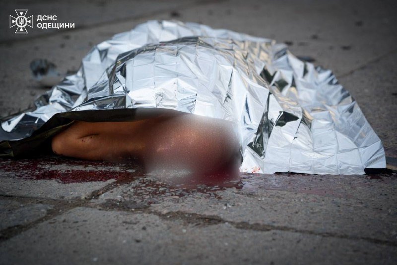 A body partially covered by a silver emergency blanket lies on paved ground with blood pooling nearby, bearing a watermark reading "ДСНС ОДЕЩИНИ" in the upper left.