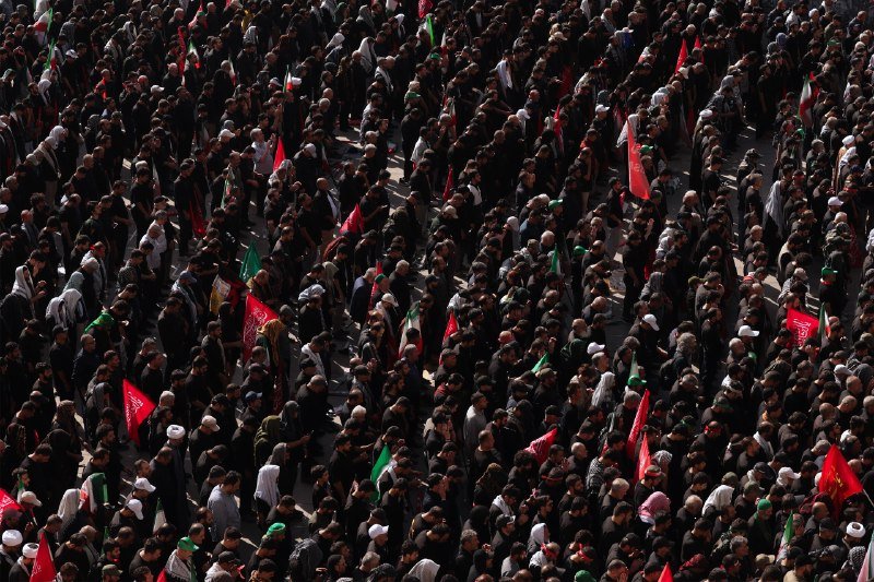 A massive crowd of people, many dressed in black, fills a public space, holding red and green flags.