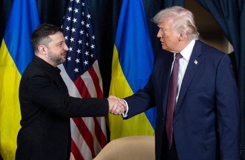 Two men in suits shake hands in front of American and Ukrainian flags.