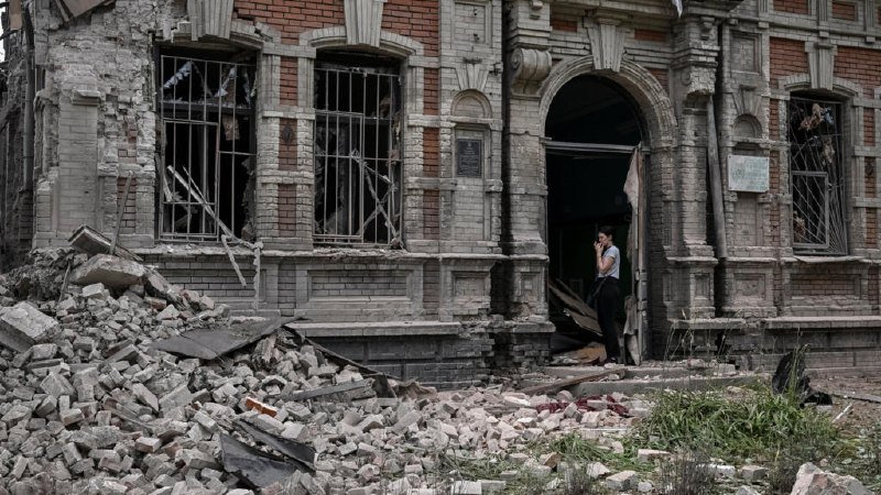 A man stands in the doorway of a heavily damaged brick building, surrounded by piles of rubble and shattered windows.