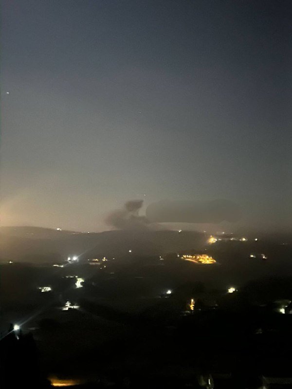 Nighttime view over a dark valley with scattered glowing lights and an illuminated hilltop structure under a deep blue sky.