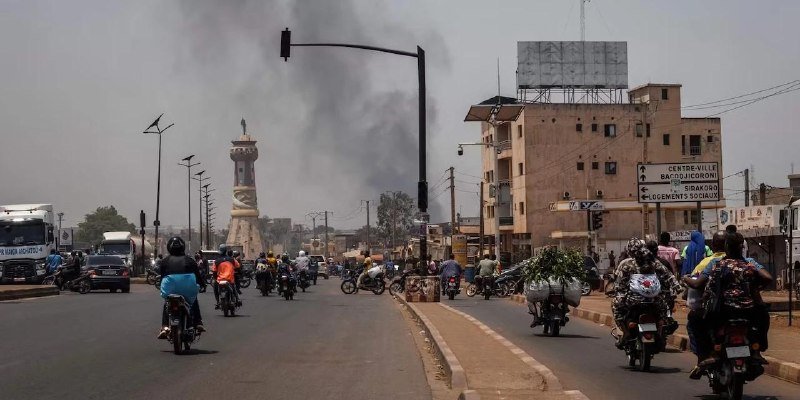 Motorcyclists and pedestrians travel along a busy urban road as a large plume of dark smoke rises behind buildings and a monument in the background.