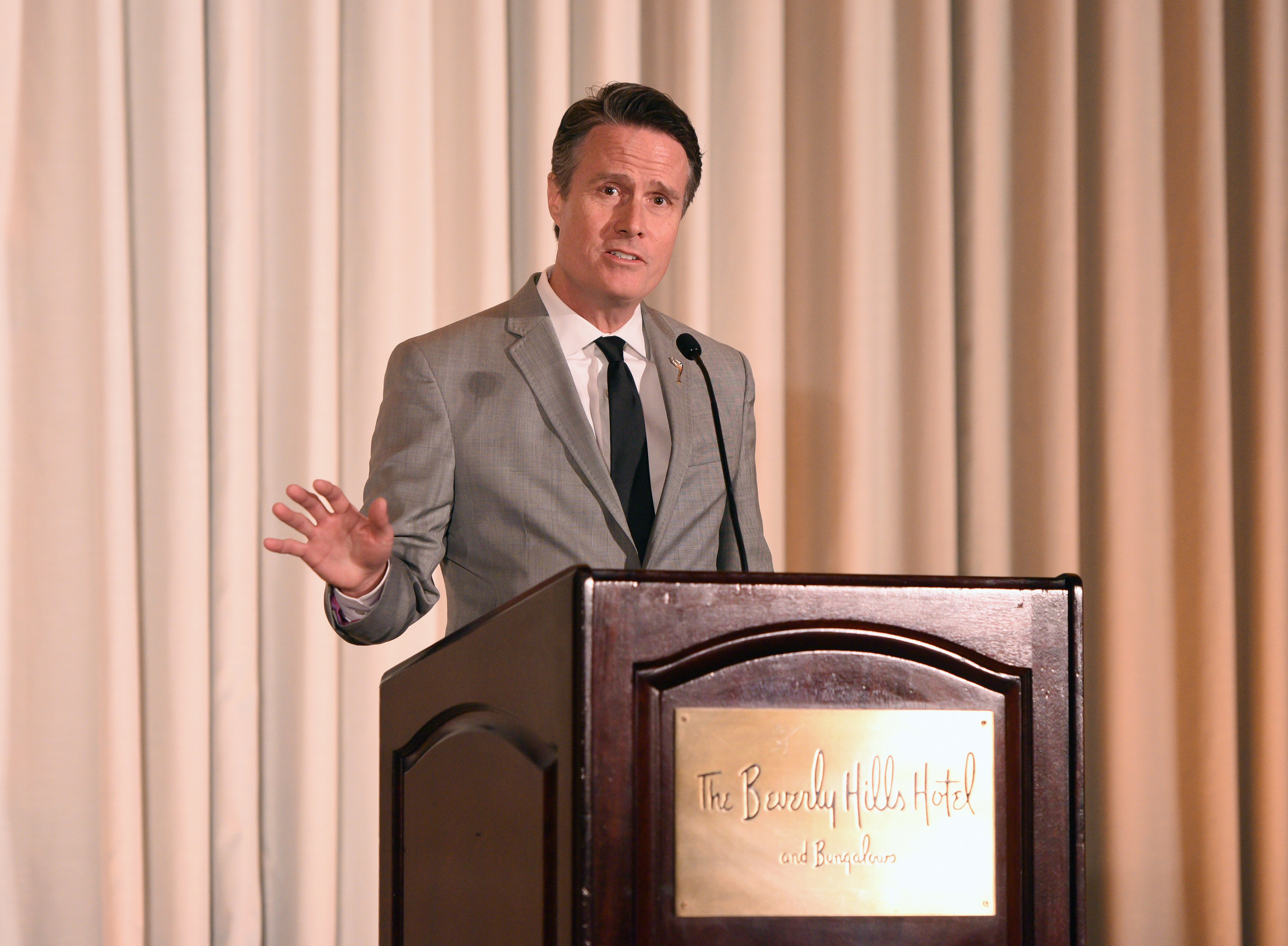 A man in a gray suit and black tie speaks at a dark wooden podium bearing "The Beverly Hills Hotel and Bungalows" placard, with cream-colored draped curtains in the background.