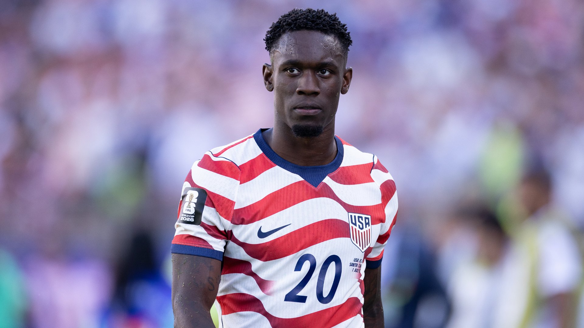 A soccer player wearing a red and white striped USA national team jersey with the number 20 stands before a blurred stadium crowd.