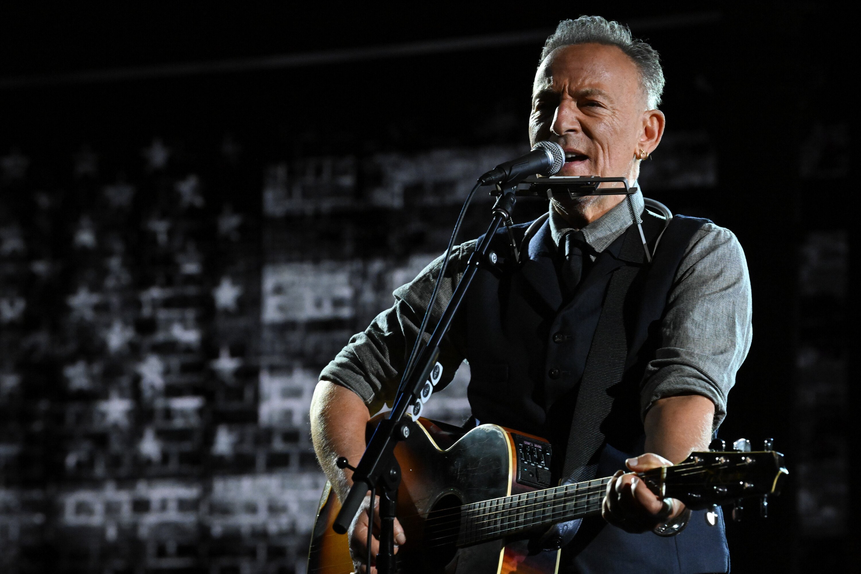 A gray-haired man in a vest and tie sings into a microphone while playing an acoustic guitar on a dimly lit stage.