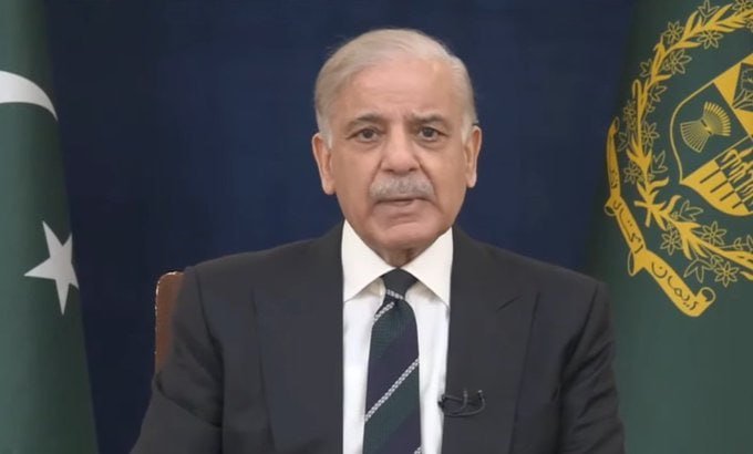 An older man with a white mustache in a dark suit speaks while seated between a green flag with a crescent and a green flag bearing a national emblem.