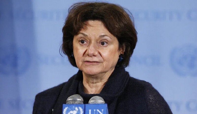 A woman with short brown hair stands at microphones marked "UN," speaking in front of a backdrop featuring a world map and partial UN insignia text.