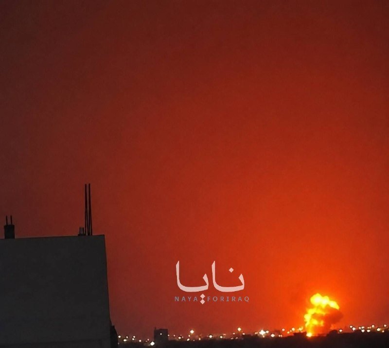 A bright explosion illuminates a dark red-orange sky behind a silhouetted rooftop, with Arabic text and "NAYA FOR IRAQ" watermark visible.