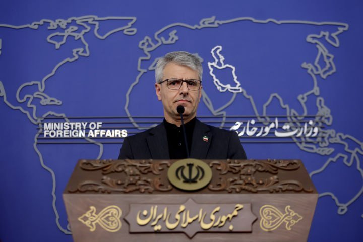 A man in a dark suit and glasses stands at an ornate podium bearing the Iranian emblem and Persian text, before a blue backdrop displaying a world map and "Ministry of Foreign Affairs."