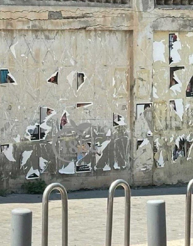 A weathered concrete wall covered in torn, peeling posters and white graffiti stands behind metal bike racks on a paved street.