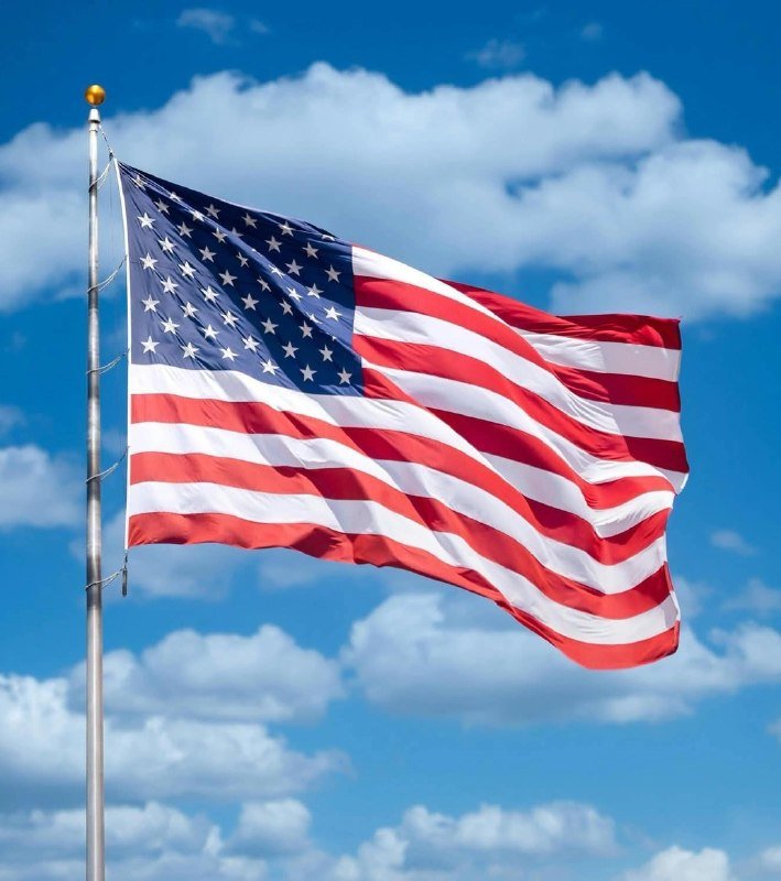 An American flag waves in the wind on a flagpole against a bright blue sky with scattered white clouds.