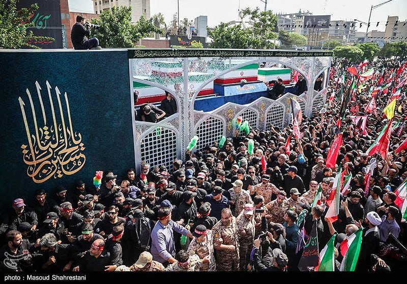A massive crowd carrying Iranian flags gathers around a decorated truck carrying a flag-draped casket in a city street, with Arabic calligraphy visible on a green banner.