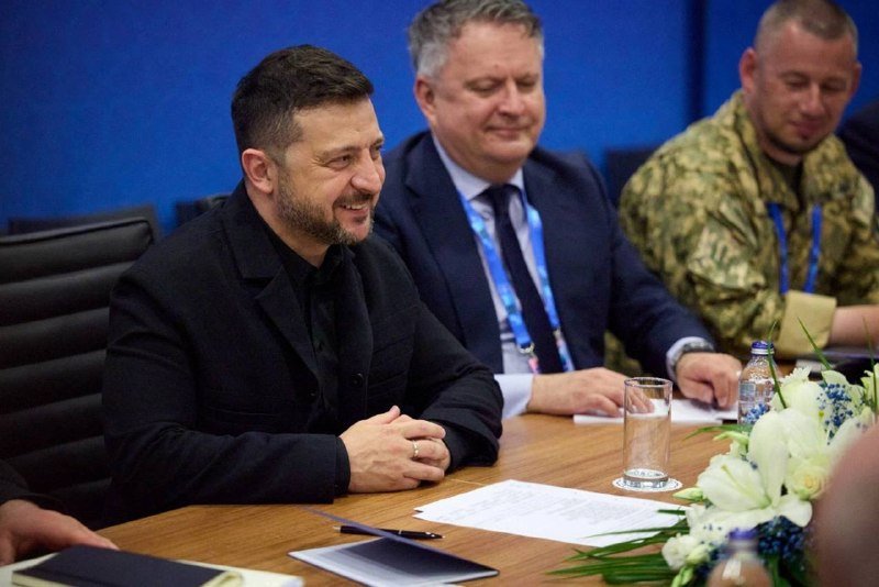 Three men sit at a conference table with documents, a water glass, and white flowers, against a blue backdrop.