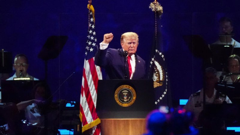 A man in a dark suit and red tie stands at a podium bearing the presidential seal, raising his right fist while speaking into a microphone.