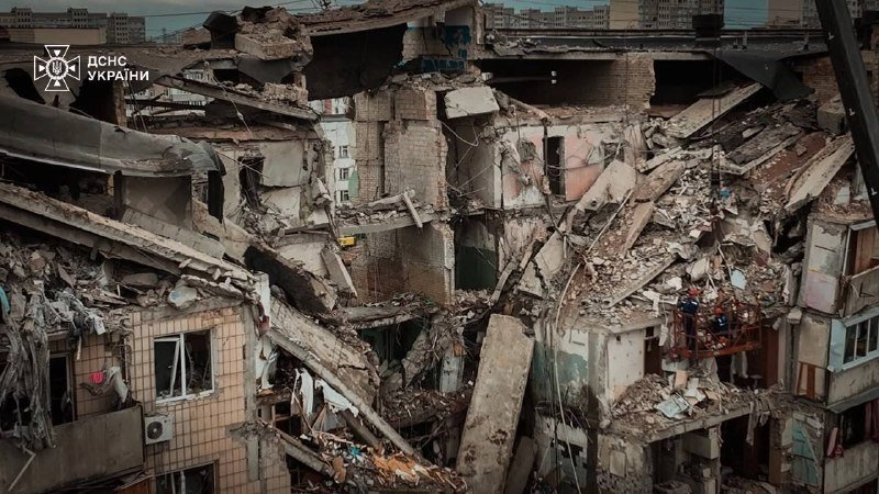 A partially collapsed multi-story residential building shows extensive structural damage, with shattered concrete, exposed rebar, and debris filling the lower floors, marked with a "ДСНС України" logo.