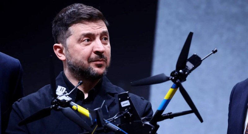 A bearded man in a black shirt stands behind a large drone with propellers painted in blue and yellow, appearing to present it at an event.