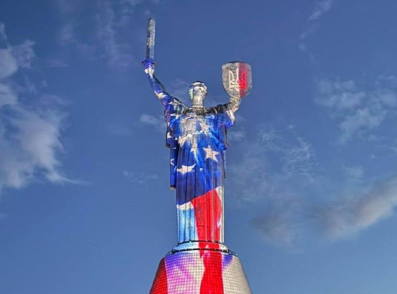 A large statue of a robed female figure holds a sword and shield aloft, illuminated in red, white, and blue against a partly cloudy sky.