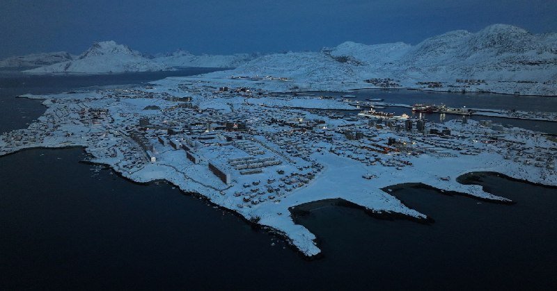 A snow-covered coastal town sits on a peninsula surrounded by dark water, with illuminated buildings and snow-capped mountains visible in the background at dusk.