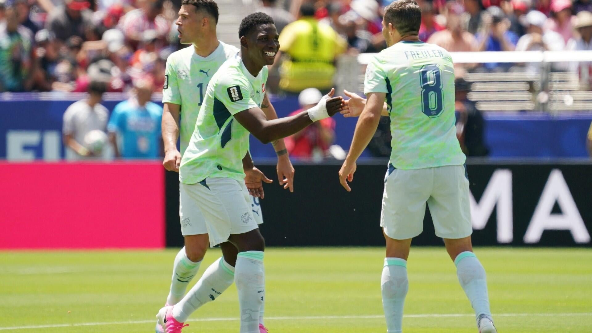 Three soccer players in white and green uniforms smile and extend their hands toward each other on a sunny pitch, with a crowded stadium visible behind them.