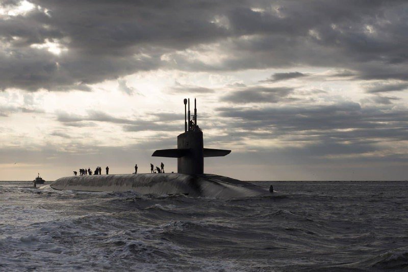 A submarine surfaces in choppy open waters under a cloudy sky, with personnel visible standing along its deck.