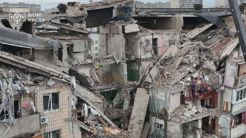 A multi-story residential building is collapsed with rubble, exposed interiors, and shattered concrete slabs scattered throughout the structure.
