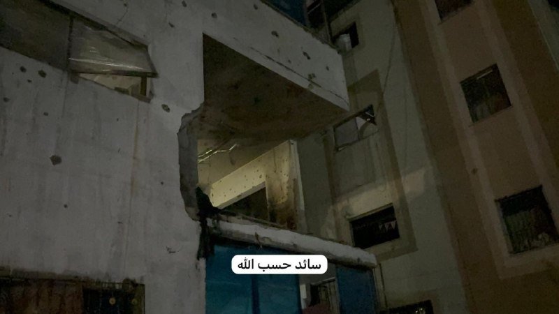 A heavily damaged multi-story building with exposed concrete, shattered openings, and Arabic text overlaid on the image.