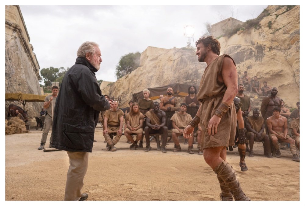 A man in a modern black coat faces a muscular man in a tan tunic and leather wrappings on a sandy set, with seated and standing onlookers watching against a rocky backdrop.