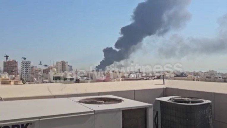 Black smoke rises from a cityscape viewed past rooftop AC units.