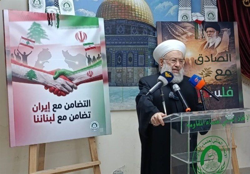 A bearded cleric in a white turban and black robe speaks at a podium with microphones, beside posters featuring flags, a handshake, and Arabic/Persian text.