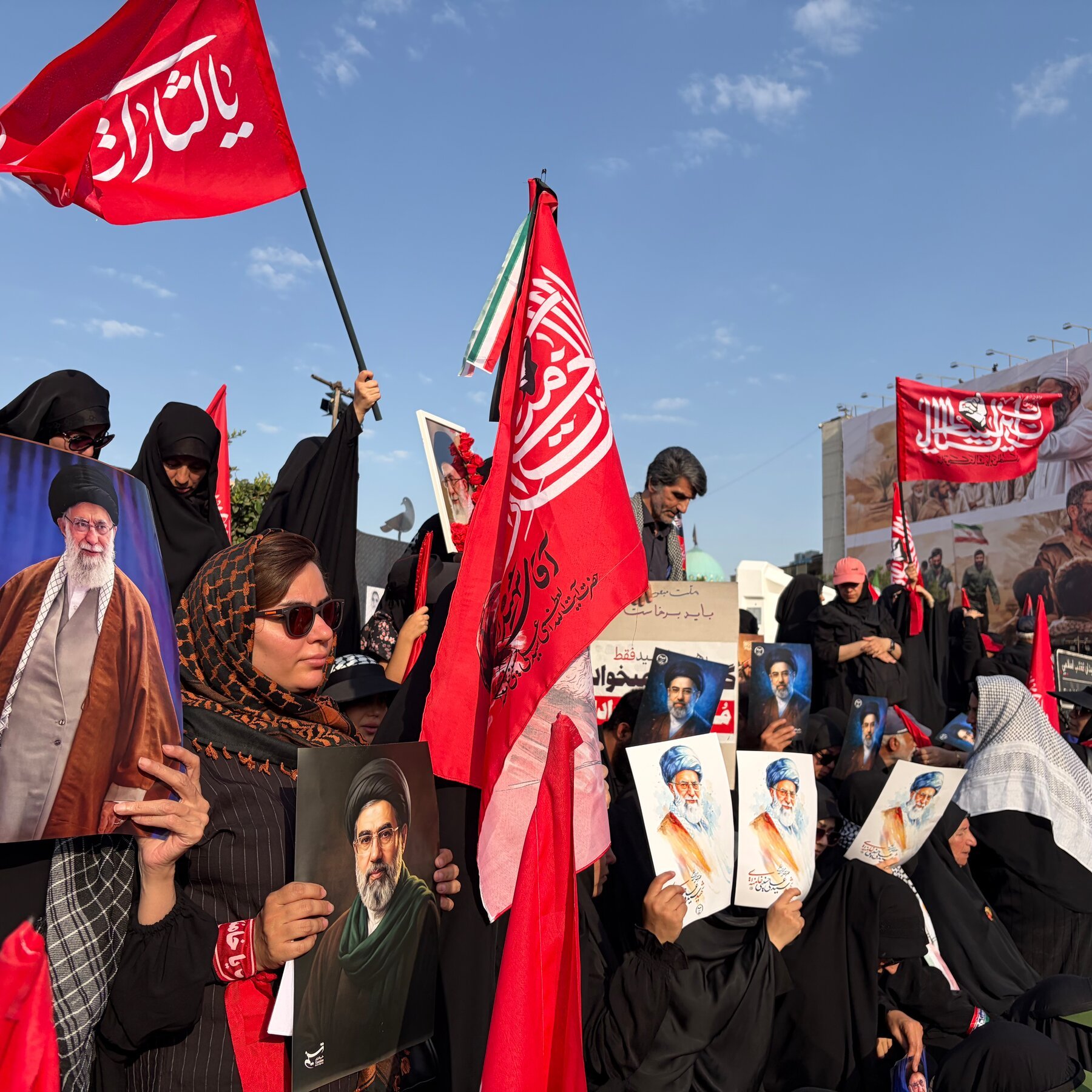 A crowd of people, many wearing black clothing, hold portraits of bearded men in turbans and red flags with Arabic text during an outdoor gathering under a partly cloudy sky.