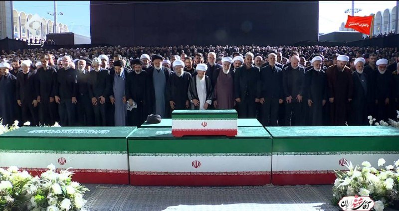 Clerics and officials in black robes stand behind three flag-draped caskets at a large outdoor funeral gathering.