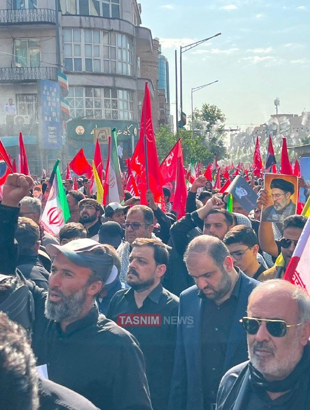 A large crowd of people marches through a city street waving red flags, Iranian flags, and portraits of a turbaned figure, with a Tasnim News watermark visible.