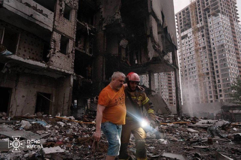 A firefighter in helmet and turnout gear supports a man in an orange shirt as they walk through rubble past a heavily damaged building, with an "ДСНС КИЄВА" logo visible.