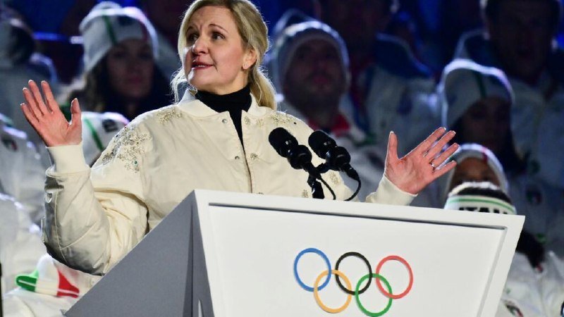 A woman in a white jacket speaks at a podium displaying the Olympic rings, gesturing with both hands raised before a crowd in white winter clothing.