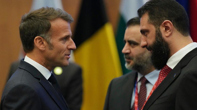 Two suited men converse face-to-face while a third bearded man stands between them, with several national flags visible in the blurred background.