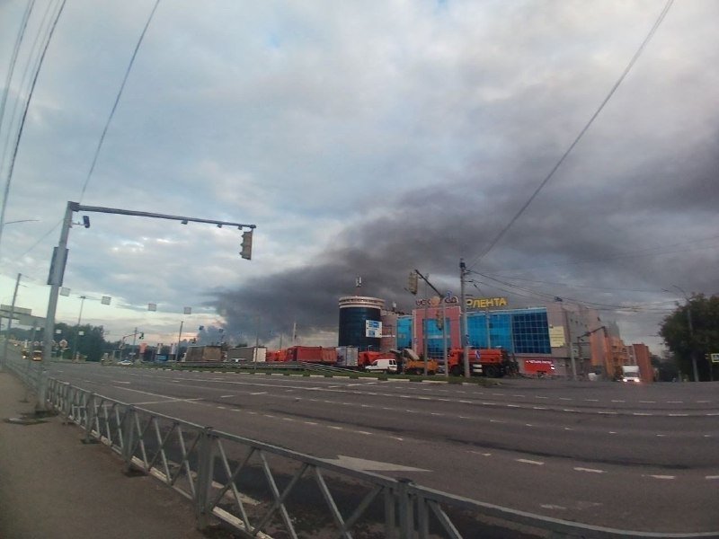 A large plume of dark smoke rises behind a commercial building and shopping center marked "Лента" along a multi-lane urban road.