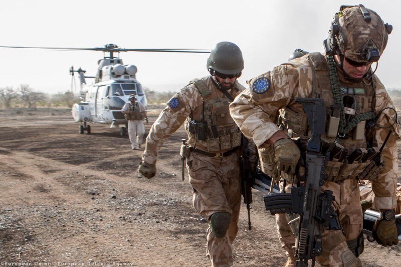 Two soldiers in desert camouflage uniforms and helmets, armed with rifles, run across a dusty landing zone toward a parked military helicopter.