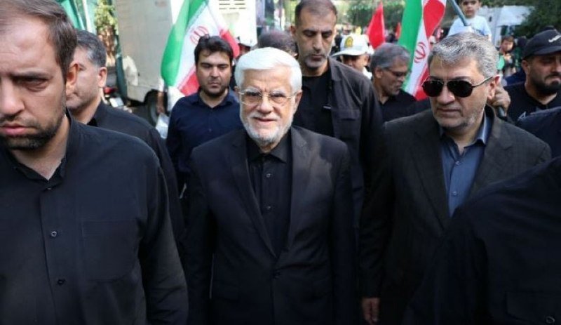 A white-bearded man in a dark suit walks through a crowd of men, with Iranian flags visible in the background.