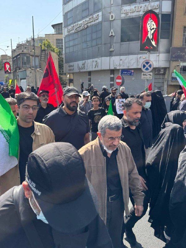 A crowd of people, including men in black and women in chadors, march on a street carrying red, green, and black flags past buildings with closed shops.