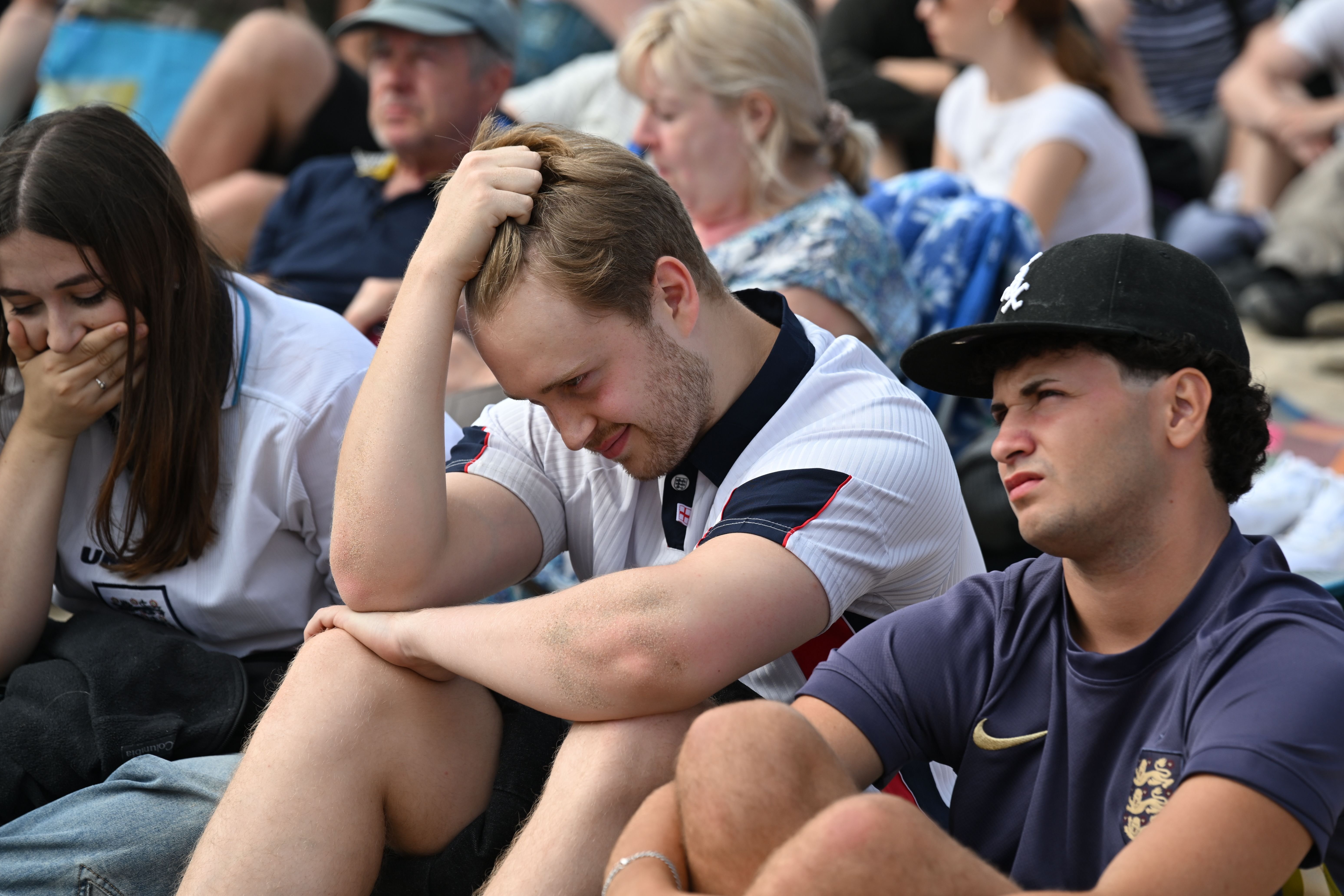 Dejected soccer fans wearing England and France jerseys sit on the ground with downcast expressions among a crowd.