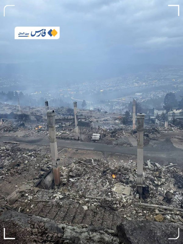 Aerial view of a burned residential neighborhood with collapsed structures, bare concrete chimneys standing amid ash and debris, and smoke visible in the distance under overcast skies.
