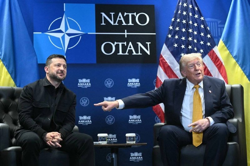 Two men in suits sit in armchairs at a NATO-branded backdrop featuring Ukrainian and American flags, with one man gesturing toward the other.