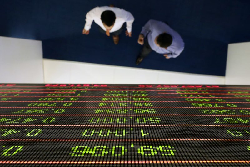 Two businessmen viewed from above stand before a large digital stock ticker displaying red and green market data on a tiled floor.
