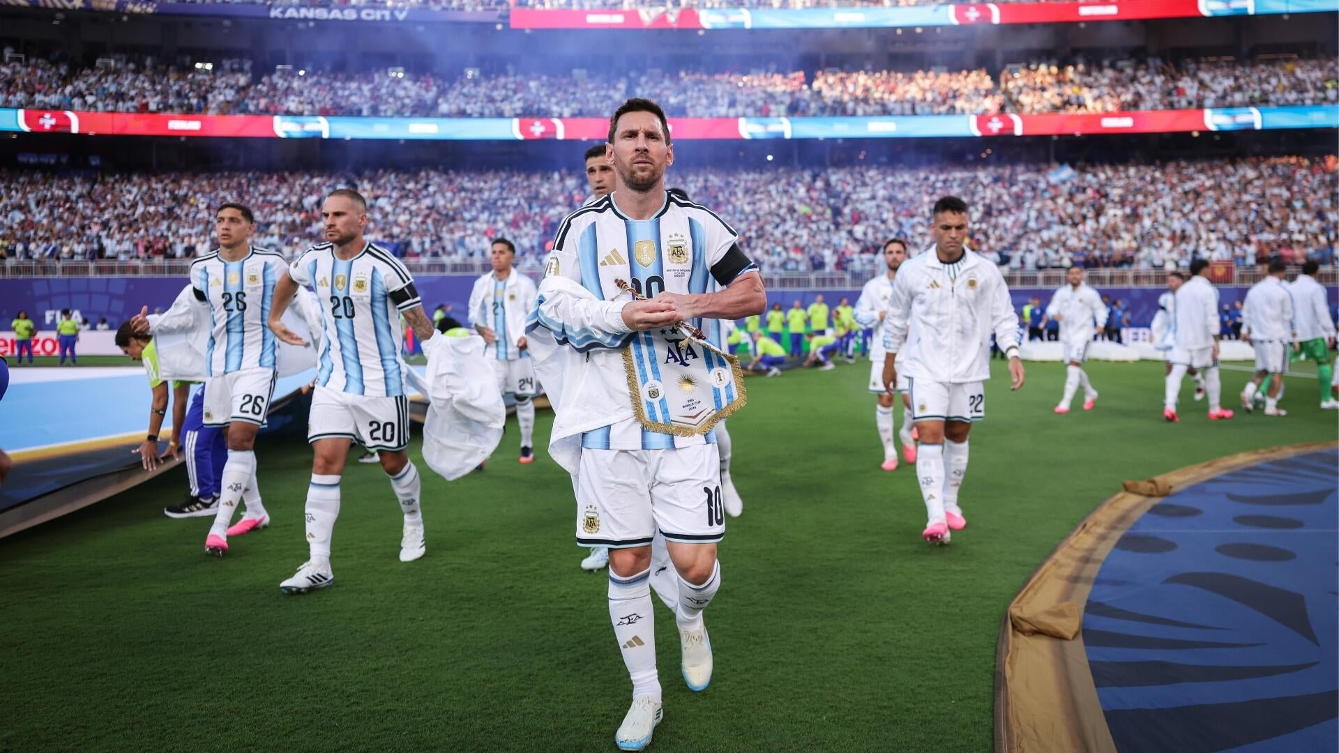 Players in light blue and white striped jerseys walk onto a soccer field in a packed stadium, led by a player carrying a small flag.