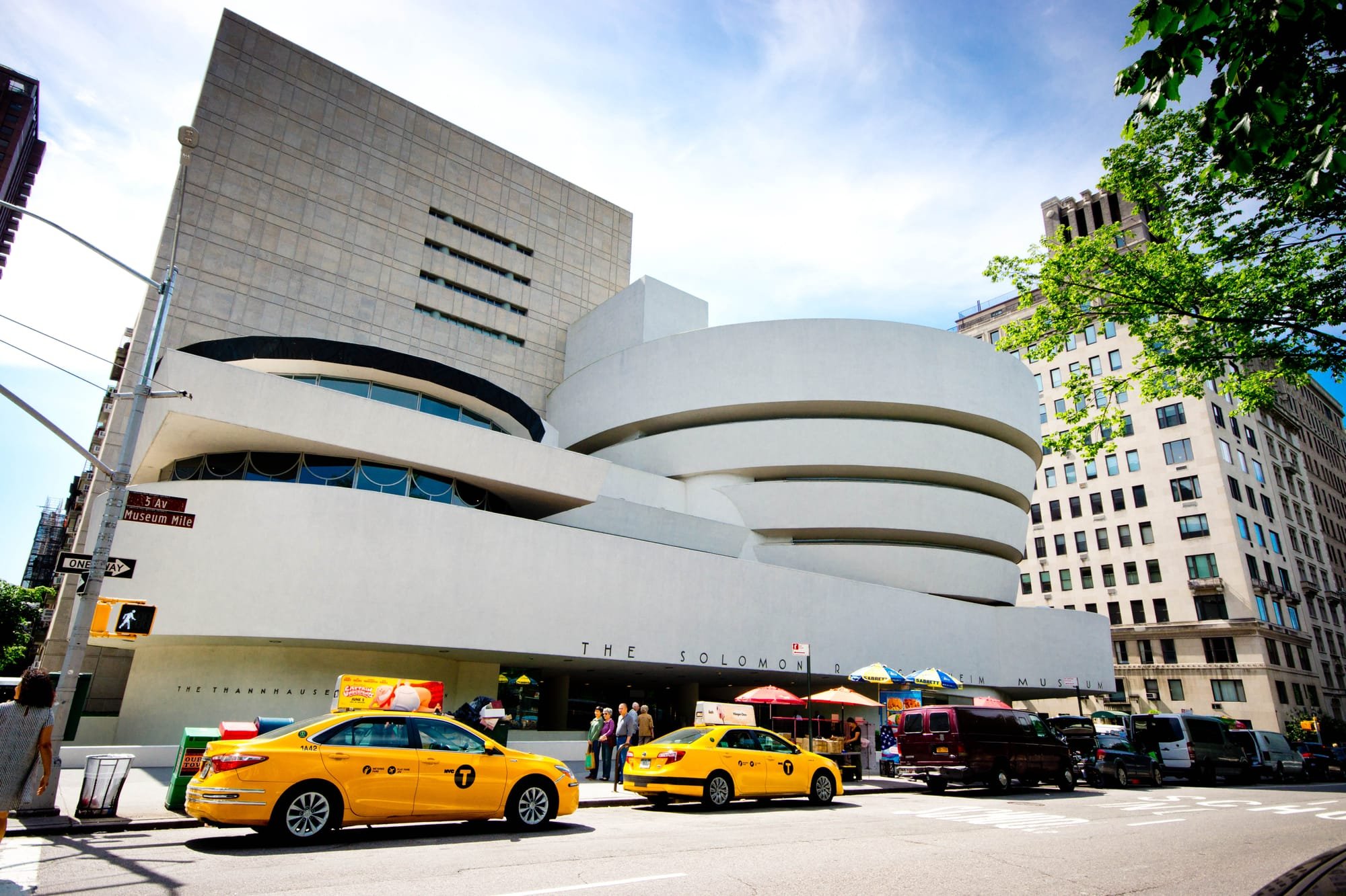 The Solomon R. Guggenheim Museum's white spiral-ramped facade stands along a busy New York City street, with yellow taxis and a SABRETT umbrella visible outside.